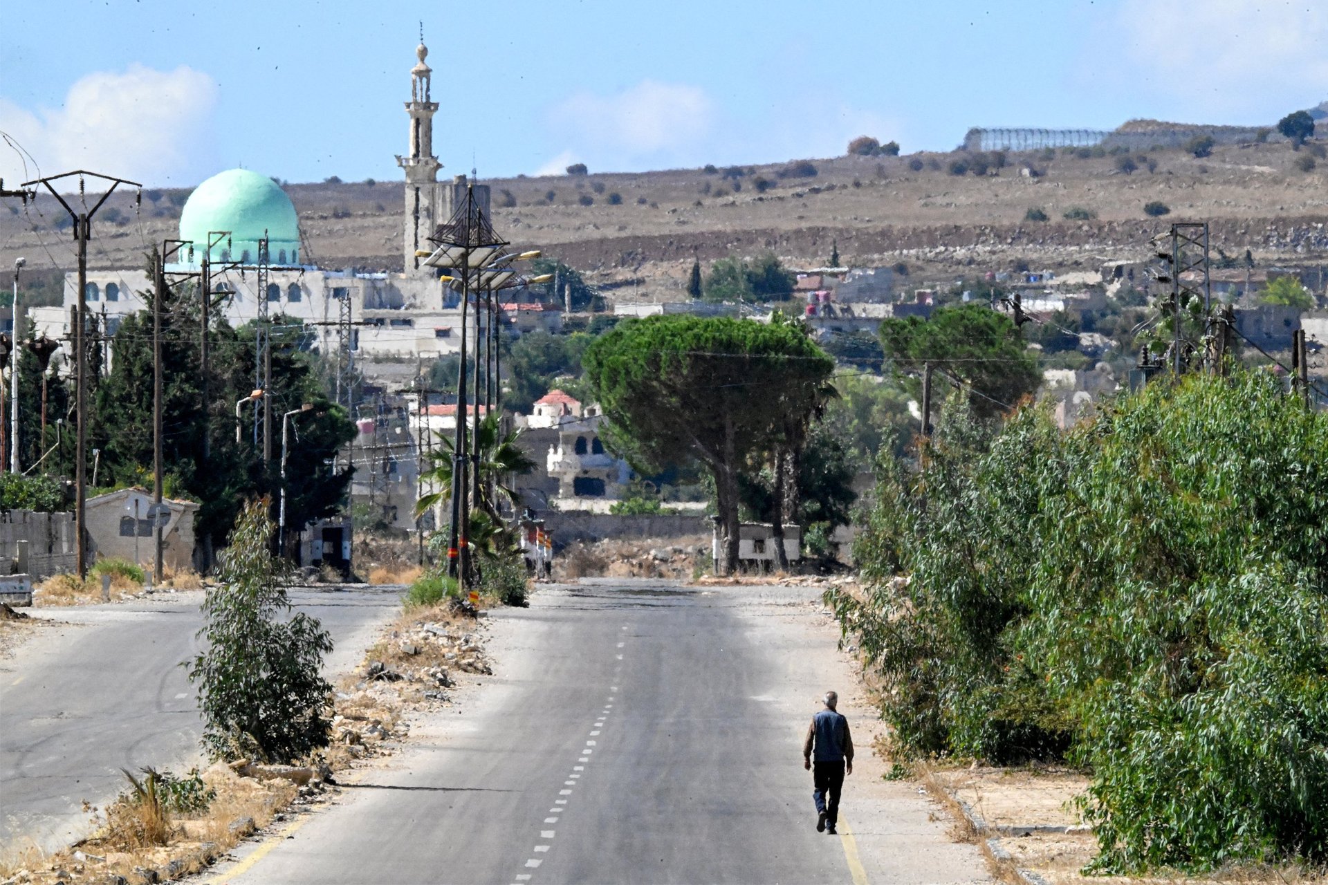 A man walks alone along an empty road towards a town with a mosque, damaged buildings and dry hills beyond